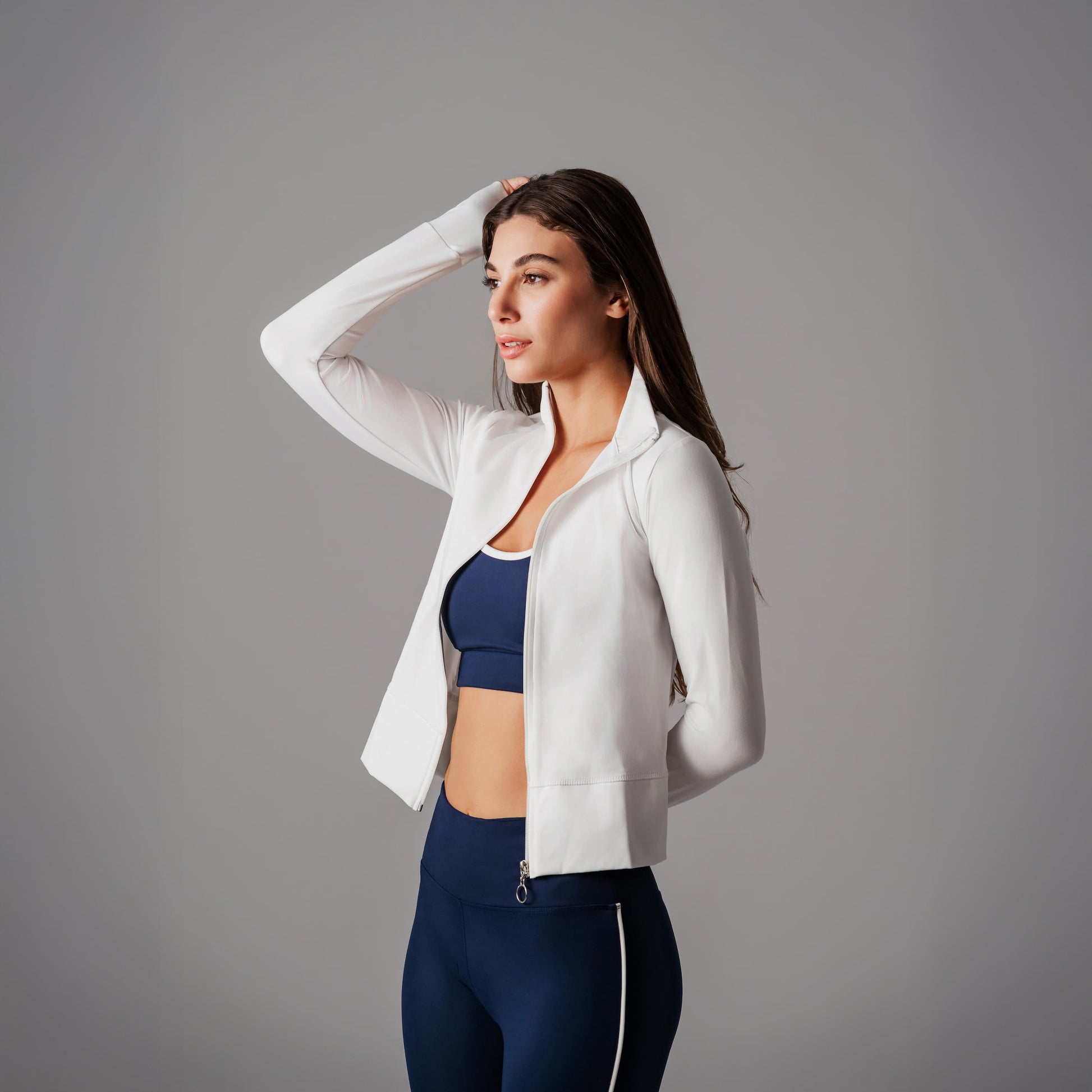 Woman wearing a white jacket over a blue sports bra and leggings on a gray background