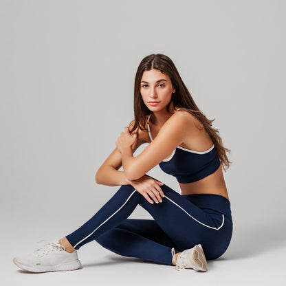 Woman wearing a navy sports bra and leggings with white accents, sitting on a light gray background.
