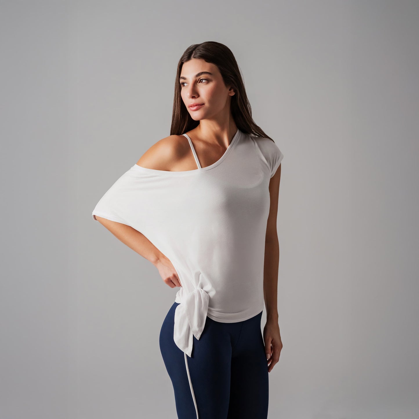 Woman wearing a white off-shoulder top with a tie detail and dark blue leggings on a gray background
