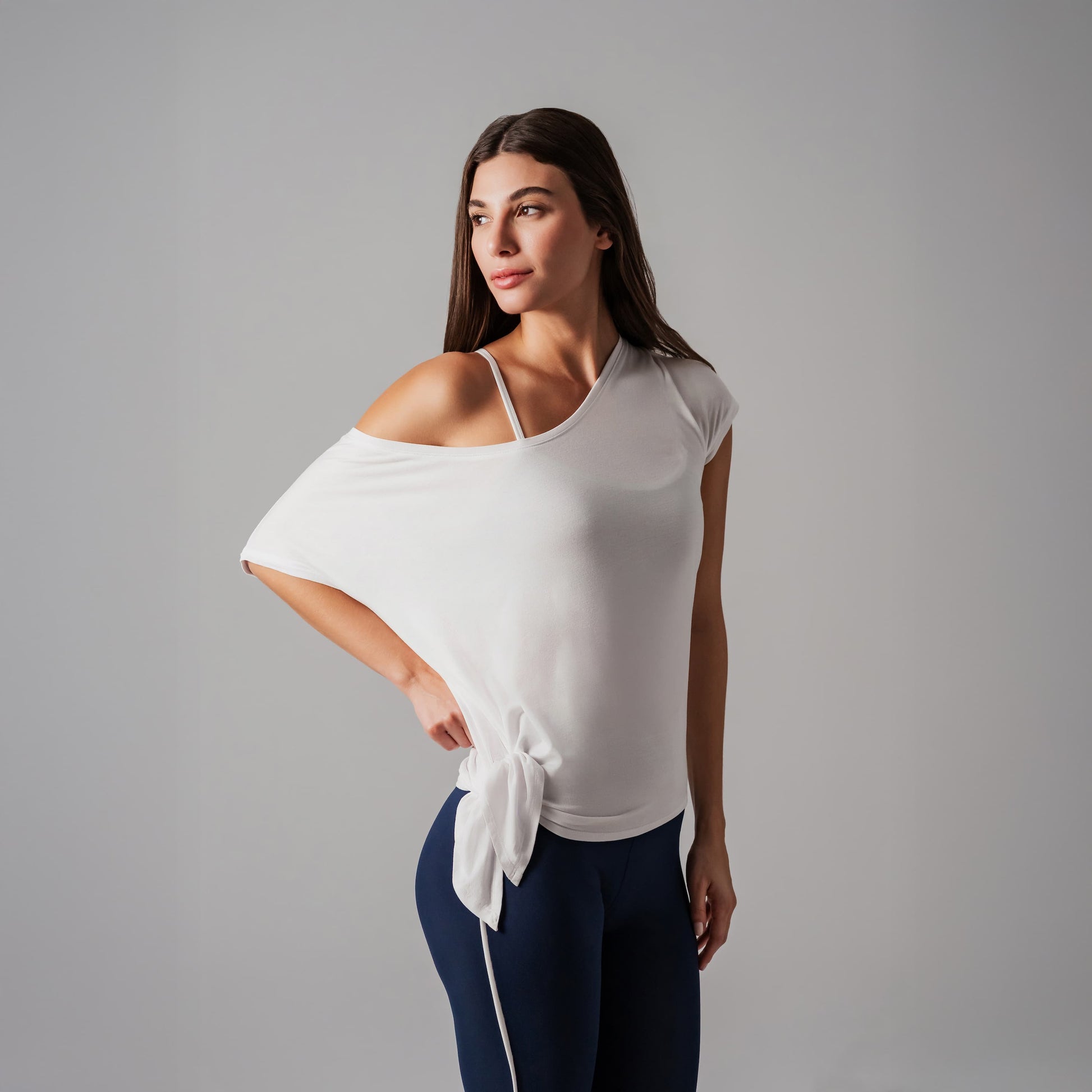Woman wearing a white off-shoulder top with a tie detail and dark blue leggings on a gray background