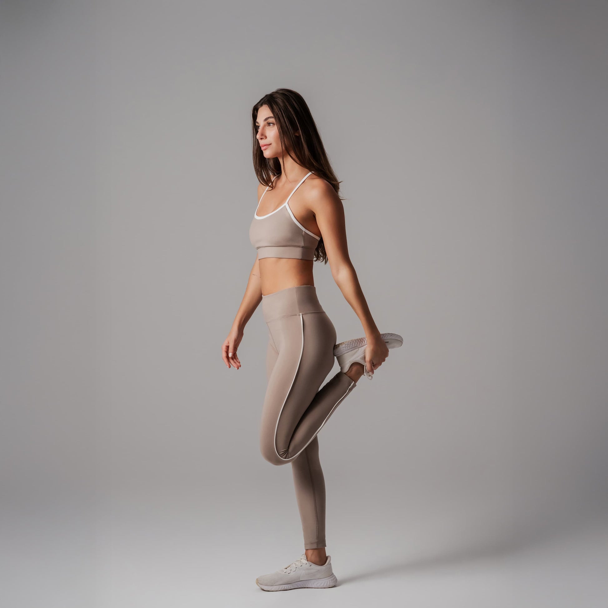 Woman in beige sports bra and leggings stretching on a gray background