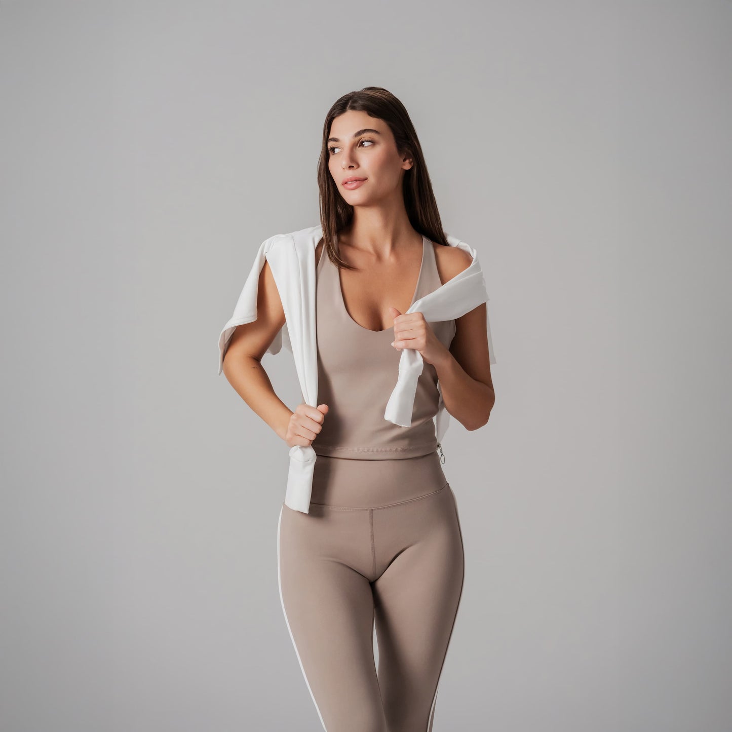Woman in beige tank top and leggings holding a white jacket against a gray background