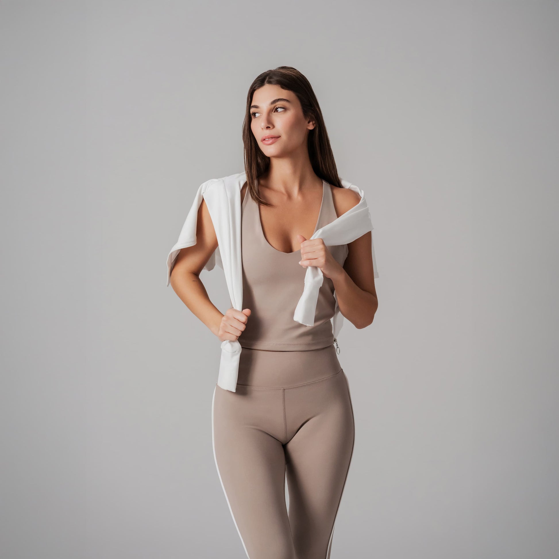 Woman in beige tank top and leggings holding a white jacket against a gray background