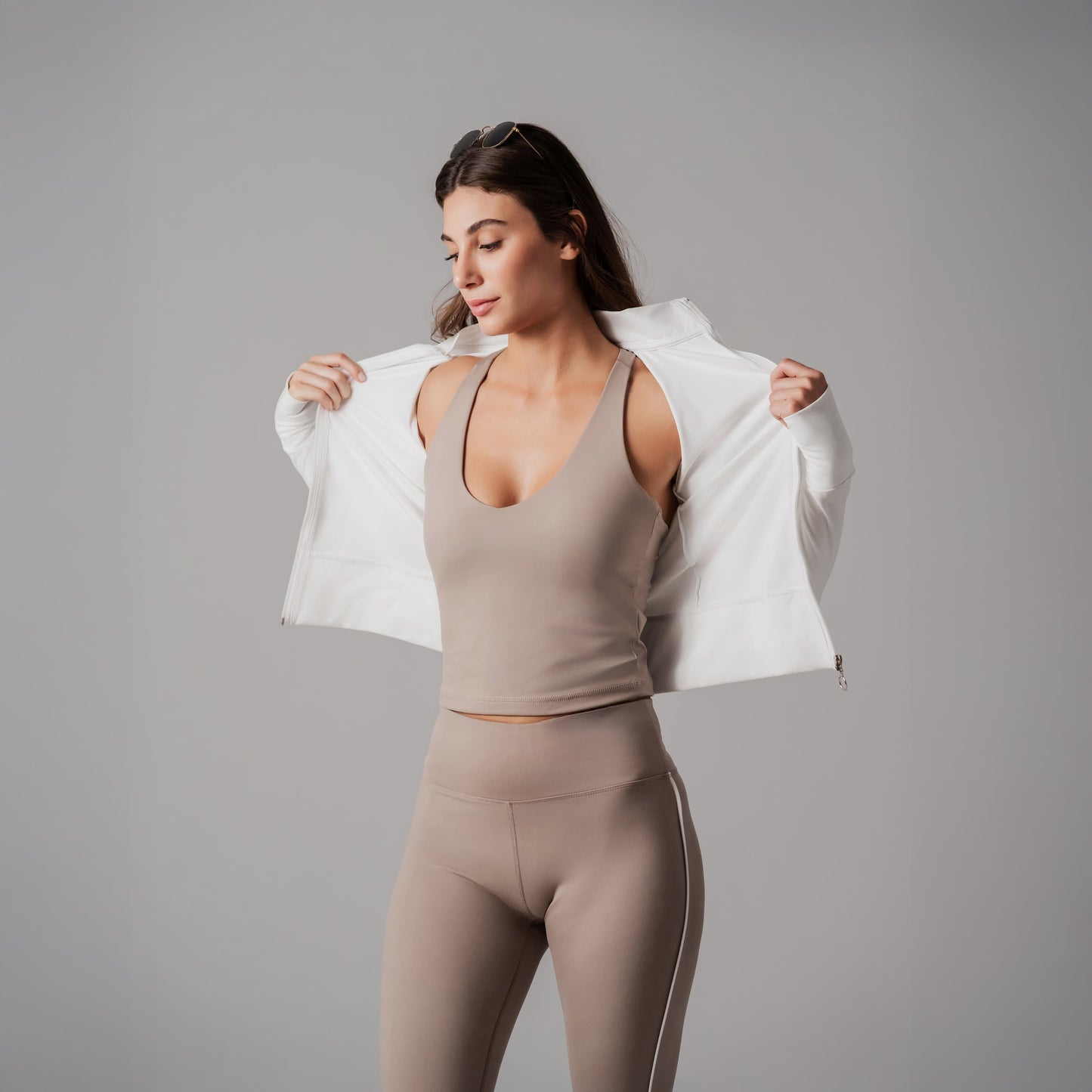 Woman wearing a beige tank top and leggings with a white jacket over her shoulders on a gray background