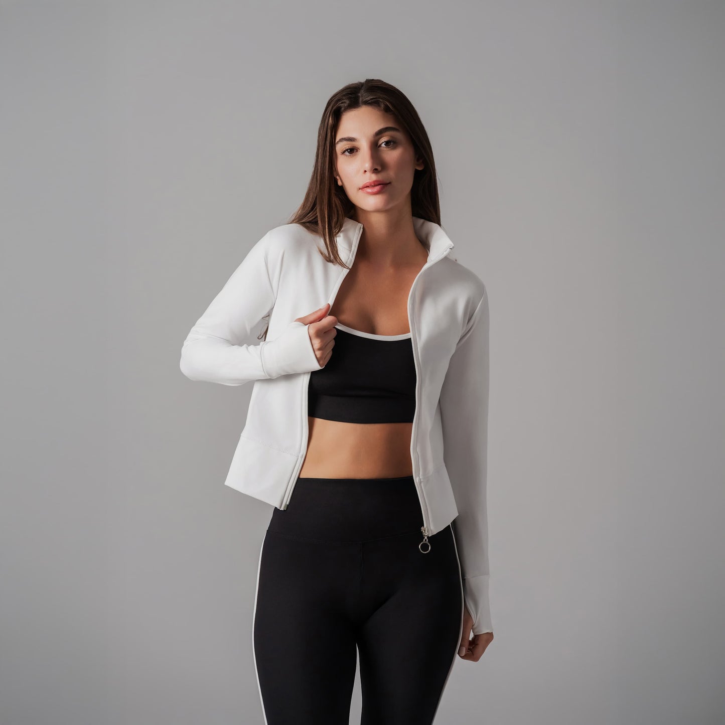 Woman wearing a white jacket over a black sports bra and leggings on a gray background