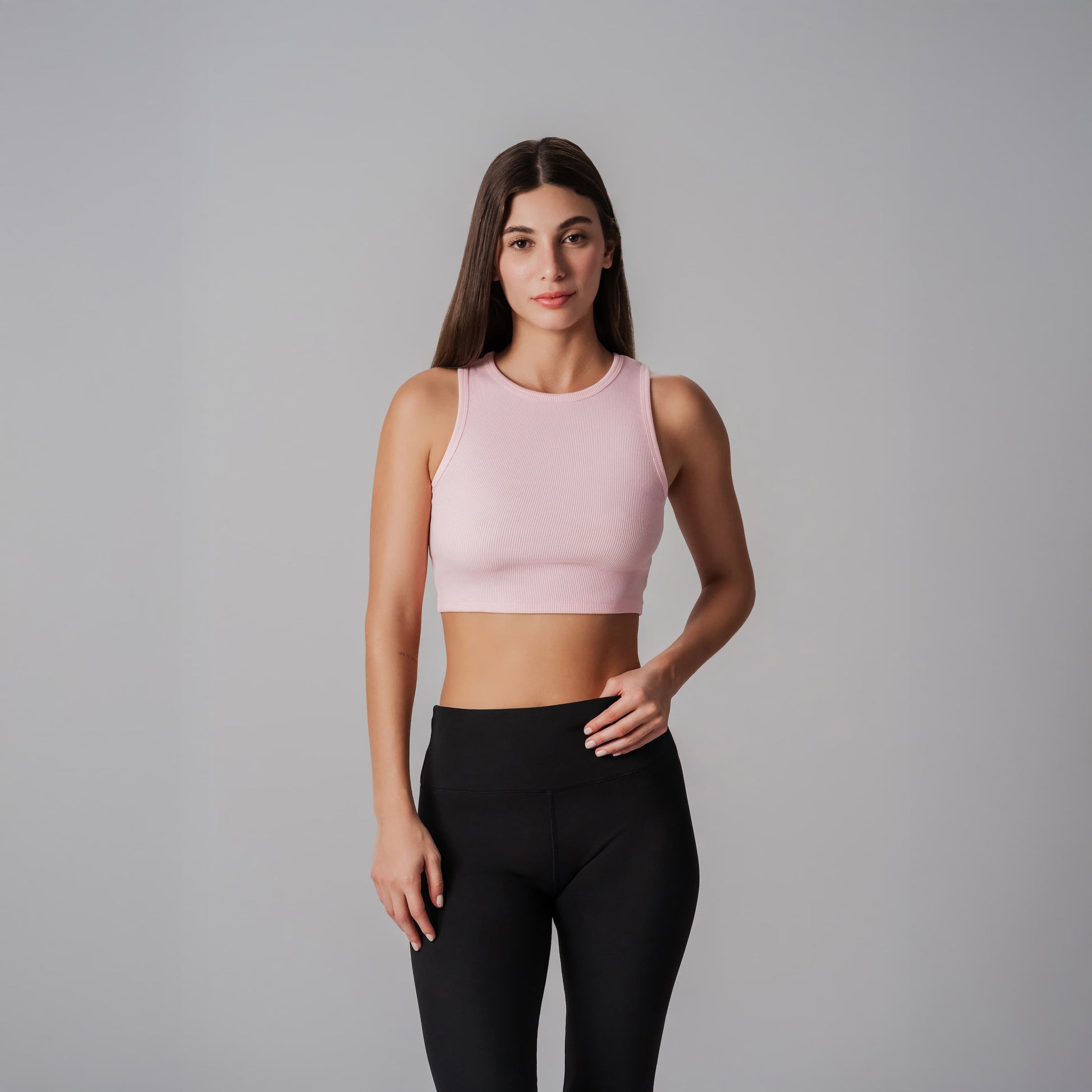 Woman wearing a pink cropped ribbed tank and black leggings on a gray background