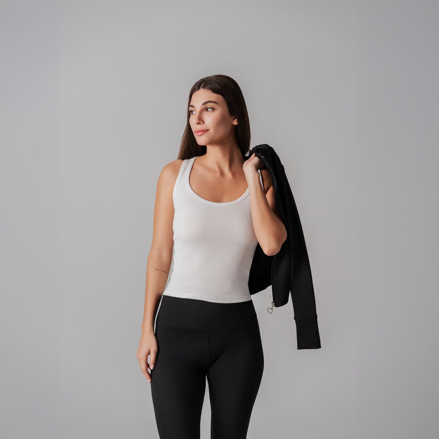 Woman wearing a white tank top and black leggings holding a black jacket against a gray background