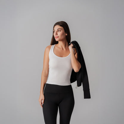 Woman wearing a white tank top and black leggings holding a black jacket against a gray background
