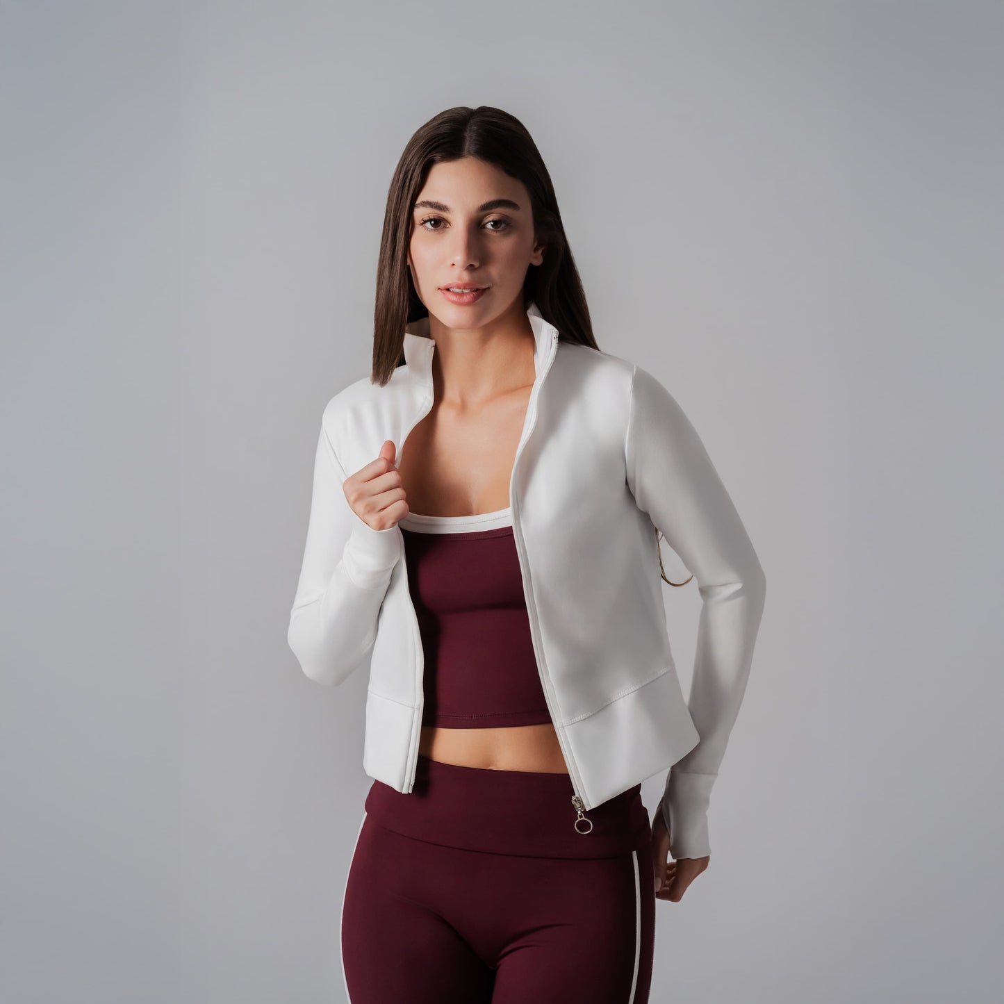 Woman wearing a white jacket over a maroon sports bra and leggings on a gray background